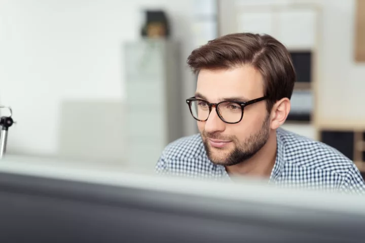 Smiling Young Businessman with Eyeglasses Sitting at his Desk and Facing at the Computer Screen, Captured in Close up