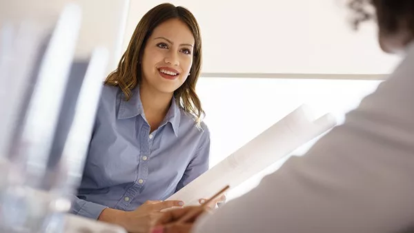 Team of two female architects, sitting at desk in office. The women talk and smile reviewing a building plan.